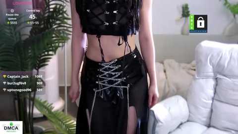 Media: Video of a fair-skinned woman with long black hair in a black lace-up corset and sheer skirt, standing in a modern living room with a white couch and potted ferns.