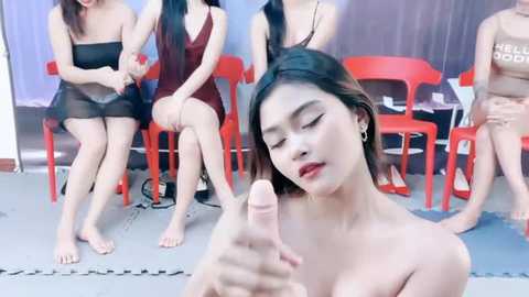 Media: Video of an Asian woman with long black hair, red lipstick, and bare chest, pointing at camera, surrounded by seated women in red chairs, in a studio setting.