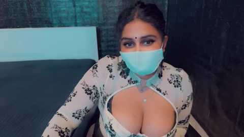 Media: Video of a South Asian woman with medium skin tone and dark hair, wearing a white floral blouse with a plunging neckline revealing cleavage, a blue face mask, and a bindi on her forehead.