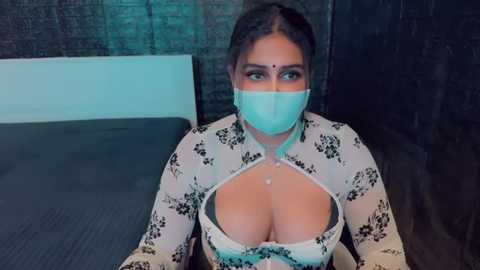 Media: Video of a curvaceous woman with medium skin tone, wearing a white floral dress with a plunging neckline, revealing ample cleavage, a face mask, and a bindi, seated indoors with dark brick walls in the background.