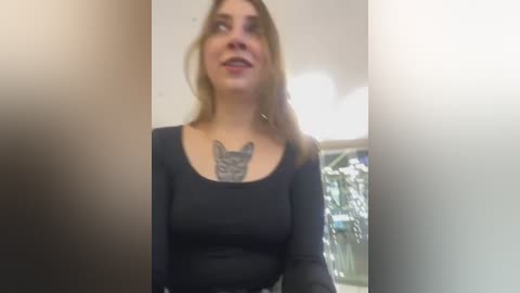 Media: Video of a young woman with fair skin and light brown hair, wearing a black long-sleeve top, showcasing a detailed cat tattoo on her chest. Background shows a blurred indoor setting with white walls and a window.