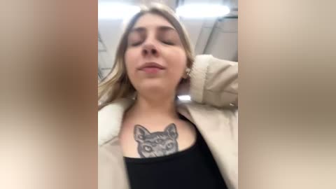 Media: A video of a young woman with light skin and blonde hair, wearing a beige jacket, displaying a black-and-grey Chihuahua tattoo on her chest. She appears relaxed, with eyes closed and a slight smile, taken in a bright, indoor setting.