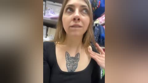 Media: A video of a young woman with shoulder-length brown hair, wearing a black top, and a large cat tattoo on her chest, standing in a brightly lit store with shelves and hanging clothes in the background.