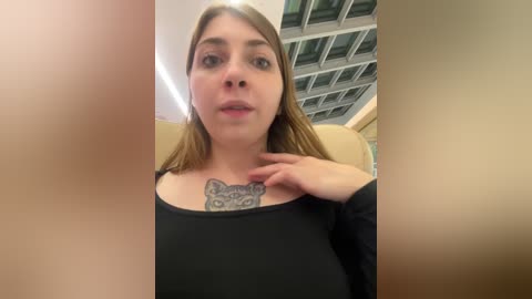 Media: Video of a young Caucasian woman with light skin, shoulder-length brown hair, and a black long-sleeve shirt. She has a detailed cat tattoo on her chest and is sitting in a modern, glass-walled room with a grid-patterned ceiling.