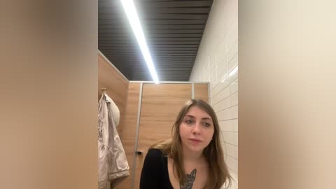 Media: Video of a young woman with long brown hair and a tattoo on her chest, standing in a narrow, brightly lit restroom with wooden stalls, white-tiled walls, and a black ceiling.