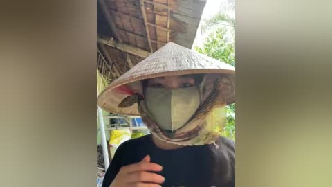 Media: Video of a person wearing a traditional conical straw hat, face mask, and black shirt, standing under a thatched-roof structure in a rural setting with greenery.