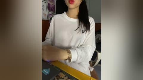 Media: Video of an Asian woman with long black hair and red lipstick, wearing a white long-sleeve shirt and gold bracelets, seated at a desk. Background includes a partially visible computer monitor and a wall with framed videos.