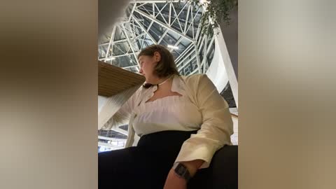 Media: Video of a woman with short light brown hair, wearing a white blouse and black skirt, seated in a modern, glass-encased atrium with a steel-framed ceiling.