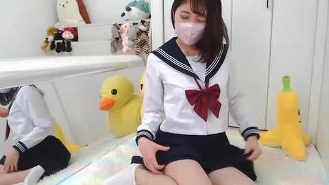 Media: Video of an East Asian woman in a white schoolgirl uniform with a red bow, sitting on a plush floor, surrounded by yellow duck toys and a mirror.