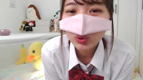 Media: Video of a young Asian woman in a white shirt, red plaid tie, and pink mask, making a kissy face, surrounded by plush toys in a white room.