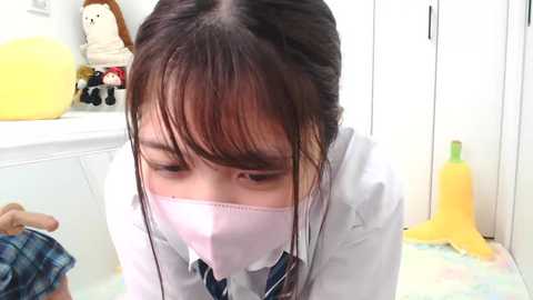 Media: Video of an Asian woman in a white lab coat and pink face mask, kneeling on a colorful mat, with a stuffed toy and a yellow bottle in the background.