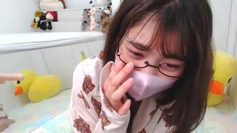 Media: Video of an Asian woman with long black hair, wearing glasses and a mask, kneeling in a bathtub filled with yellow rubber duckies, white floral pajamas, and white walls.