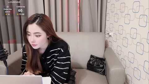 Media: Video of a young woman with long brown hair, wearing a striped sweater, sitting on a beige couch in a modern room with patterned wallpaper. She's using a laptop, and a black handbag is on the couch.