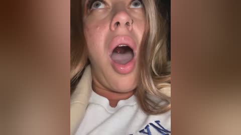 Media: Video of a shocked young woman with light skin, blue eyes, and blonde hair, mouth open in surprise, wearing a white T-shirt.