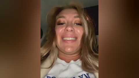 Media: Video of a smiling Caucasian woman with blonde hair, wearing a white t-shirt with blue text, in a dimly-lit room.