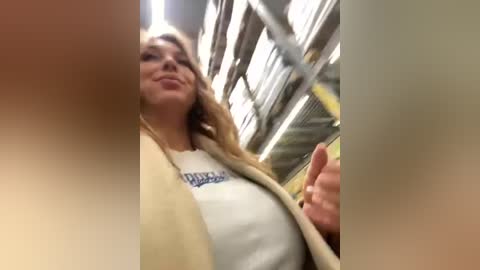 Media: Video of a young Caucasian woman with light skin and long blonde hair, wearing a beige coat and white shirt, smiling, taken from a low angle inside a dimly lit, industrial-looking room with metal beams and overhead lights.