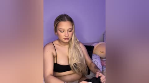 Media: Video of a blonde, fair-skinned woman with a curvy physique wearing a black bra, seated on a bed with stuffed animals. She has a tattoo on her left arm.