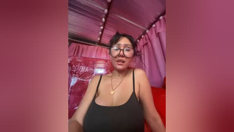 Media: A video of an Asian woman with glasses, wearing a black spaghetti-strap top, in a pink-tinted room with plastic sheets and a corrugated metal ceiling.