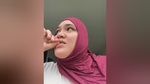 Media: Video of a young woman with a medium complexion and wearing a maroon hijab, gently touching her lips with her index finger. Background shows a blurred, muted green wall.