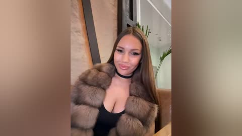 Media: Video of a young woman with long brown hair, wearing a black choker and a fur coat, seated indoors, with a blurred background featuring a plant and white walls.