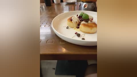 Media: Video of a round white plate with two small, golden-brown cakes topped with green microgreens and red sauce, set on a wooden table in a cozy, dimly lit cafe.