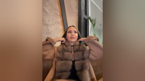 Media: Video of a Caucasian woman with light skin, long brown hair, wearing a luxurious brown fur coat and black bra, seated in a modern, minimalistic room with beige walls and a green potted plant.