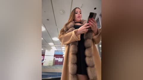 Media: Video of a light-skinned woman with long, straight brown hair, wearing a beige fur coat over a black dress, holding a smartphone in a modern indoor setting with fluorescent lights.