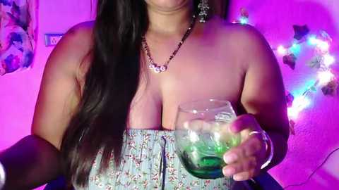 Media: Video of a light-skinned woman with long, straight black hair, wearing a strapless floral top, holding a green cocktail glass. Background features colorful string lights and a framed picture.