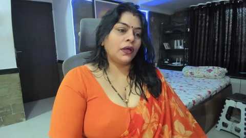 Media: Video of a woman with long black hair, wearing an orange floral sari, sitting on a gray couch in a modern bedroom with a bed and bookshelf.