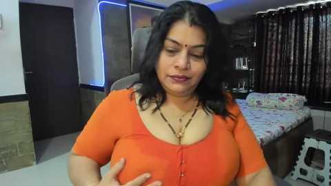 Media: Video of a curvy, middle-aged South Asian woman with medium skin tone, wearing an orange top, in a simple bedroom with floral bedspread, dark curtains, and a doorway.