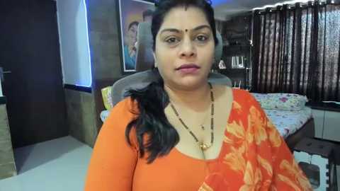 Media: Video of a South Asian woman with medium skin tone, wearing an orange sari with floral pattern, seated in a modern bedroom with dark curtains, bed, and framed portrait.