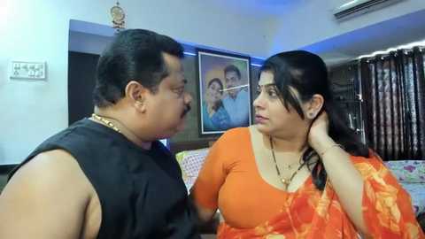 Media: Video of a middle-aged man with short hair, wearing a black sleeveless shirt, and a woman with long dark hair, dressed in an orange saree, engaged in conversation indoors, with framed portraits and curtains in the background.