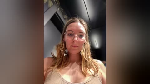 Media: Video of a young Caucasian woman with fair skin, long blonde hair, and clear glasses, wearing a beige tank top. She stands indoors against a dimly lit, industrial background.