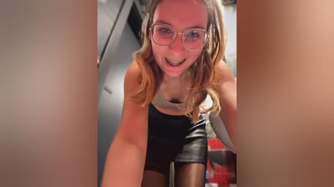 Media: Video of a young Caucasian woman with fair skin and long, wavy blonde hair, wearing glasses and a black leather mini-skirt. She's leaning forward, smiling seductively, in a dimly-lit room.