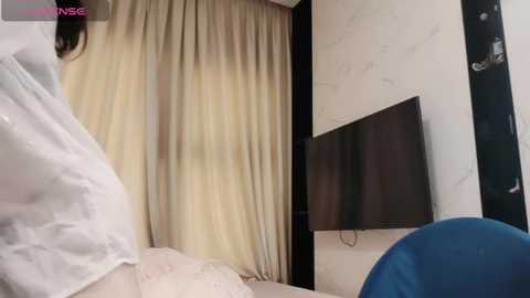 Media: Video of a modern, minimalist bedroom with beige curtains, white walls, a flat-screen TV, and a blue chair, featuring a woman in white pajamas partially visible.