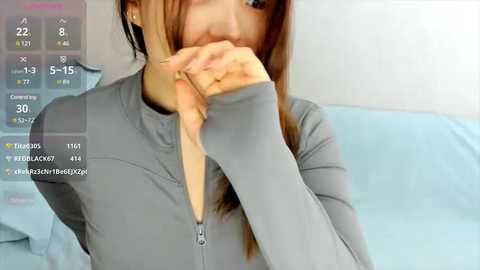 Media: A video of a young Asian woman with straight brown hair, wearing a light grey long-sleeve top, covering her mouth with her right hand. The background features a light blue bedspread and a blurred white wall. The image includes a digital clock and text overlay.