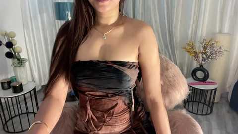 Media: Video of a woman with long brown hair, wearing a strapless, textured brown and black dress, seated in a plush chair. She wears a delicate necklace. Background features white curtains, a black cylindrical table, and a vase with dried flowers.