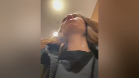 A blurry video of a woman with light skin, wearing a grey hoodie, standing with her head tilted back, possibly in a bathroom, with warm lighting and a beige wall in the background.