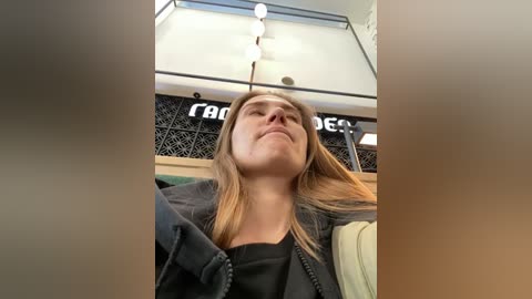 Video of a young woman with straight blonde hair, wearing a black jacket, lying back in a dimly lit room with a patterned ceiling and \"Taco Bell\" signage.