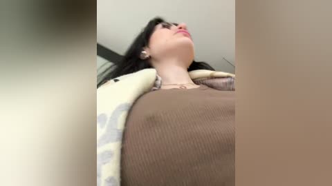 Media: Video of a young woman with long black hair, fair skin, and a slender physique, wearing a brown top and a patterned yellow and gray sweater, captured from a low angle emphasizing her cleavage.