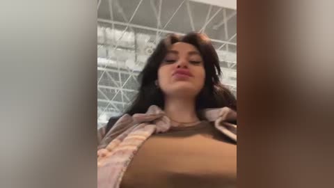 Media: Video of a young woman with long dark hair, wearing a brown shirt and plaid jacket, standing in a modern, industrial building with white beams and large windows.