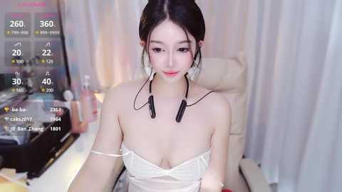 Media: Video of a young Asian woman with pale skin, dark hair in a bun, wearing a white off-shoulder top and black headphones, seated in a modern room with a computer screen displaying a live stream.