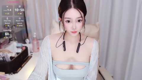 Media: Video of an Asian woman with light skin and black hair styled in pigtails, wearing a light blue top and a white cardigan, sitting in a cluttered room with makeup and skincare products on a table.