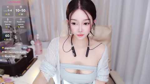 Media: Video of an East Asian woman with fair skin, black hair in a bun, wearing a light blue crop top and headphones, seated at a desk with a computer, background showing a white curtain.