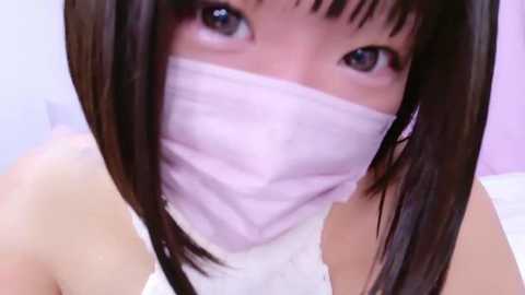 Media: Close-up video of an East Asian woman with shoulder-length straight black hair, wearing a white surgical mask, and a white sleeveless top.