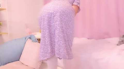 Media: Video of a person in a purple floral dress, standing in a softly lit bedroom with pastel pink walls, a bed with white sheets, and a blue cushion.