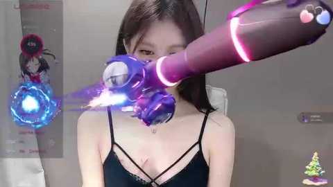 Media: Video of a young East Asian woman with straight black hair, wearing a black spaghetti-strap top, holding a futuristic purple and pink robotic arm with glowing blue lights. Background features a wall with posters and a Christmas tree ornament.