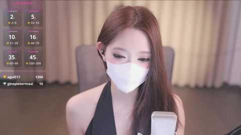 Media: Video of a young Asian woman with long brown hair, wearing a black halter top, white face mask, and a white rectangular box. She sits in a room with beige curtains and a gray chair.