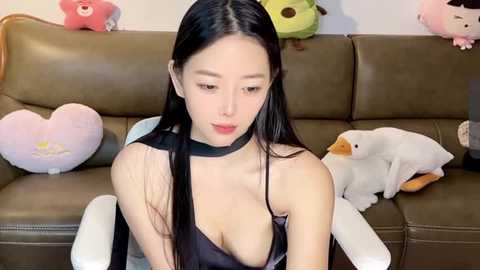 Media: Video of an Asian woman with long black hair, wearing a low-cut black dress, sitting on a brown leather couch. Background includes plush toys, including a heart-shaped pillow and a duck.