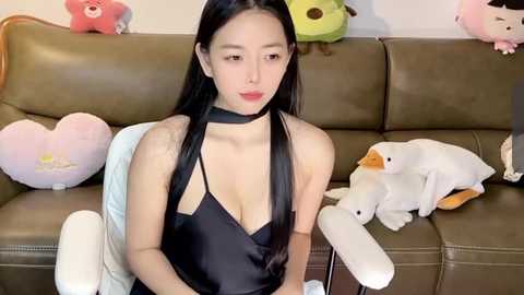 Media: Video of an Asian woman with long black hair, wearing a black halter top, sitting on a brown leather couch with plush toys.